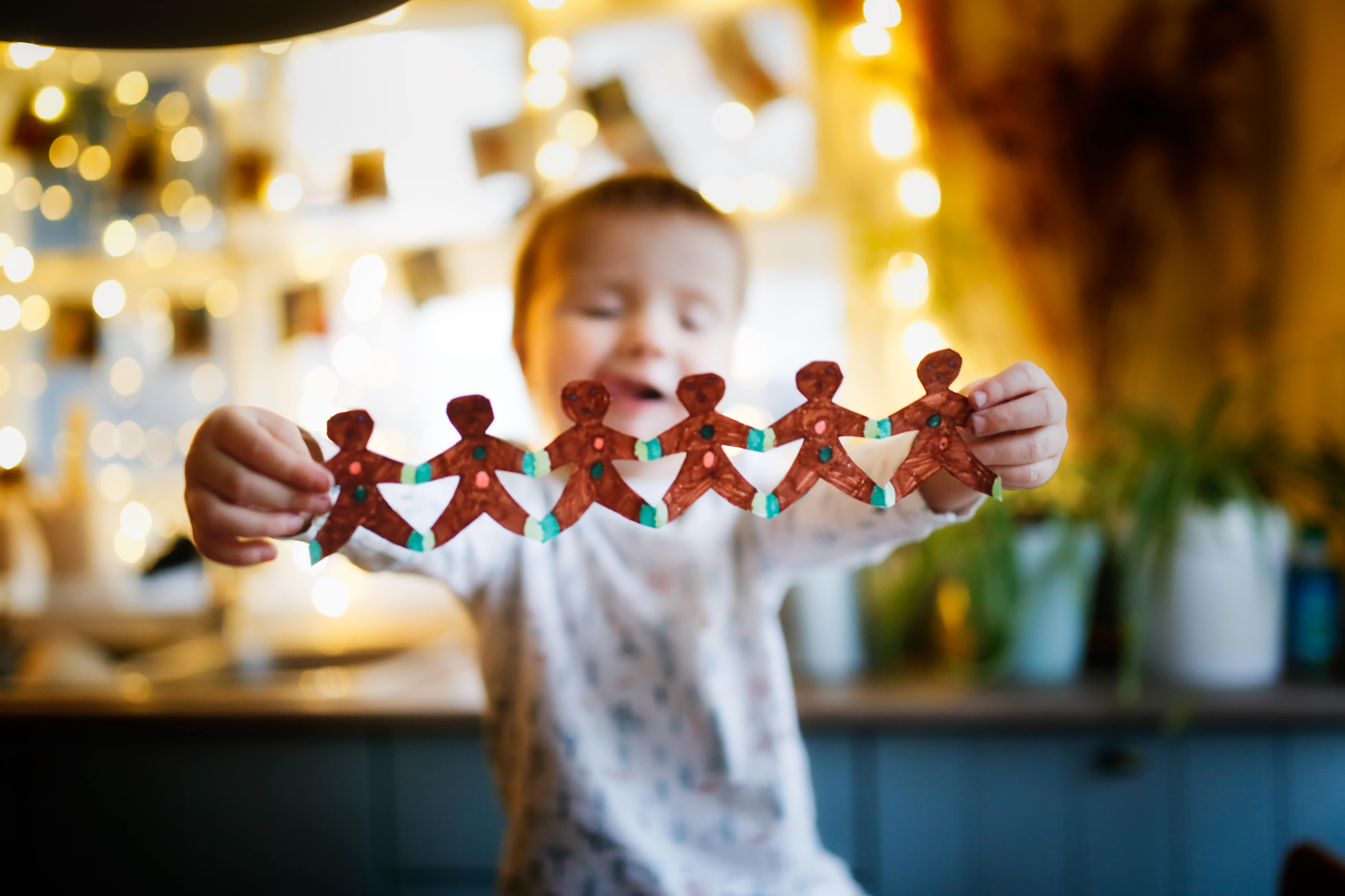 A preschool-age child holding a line of gingerbread people holding hands, showing off their Christmas craft for kids.