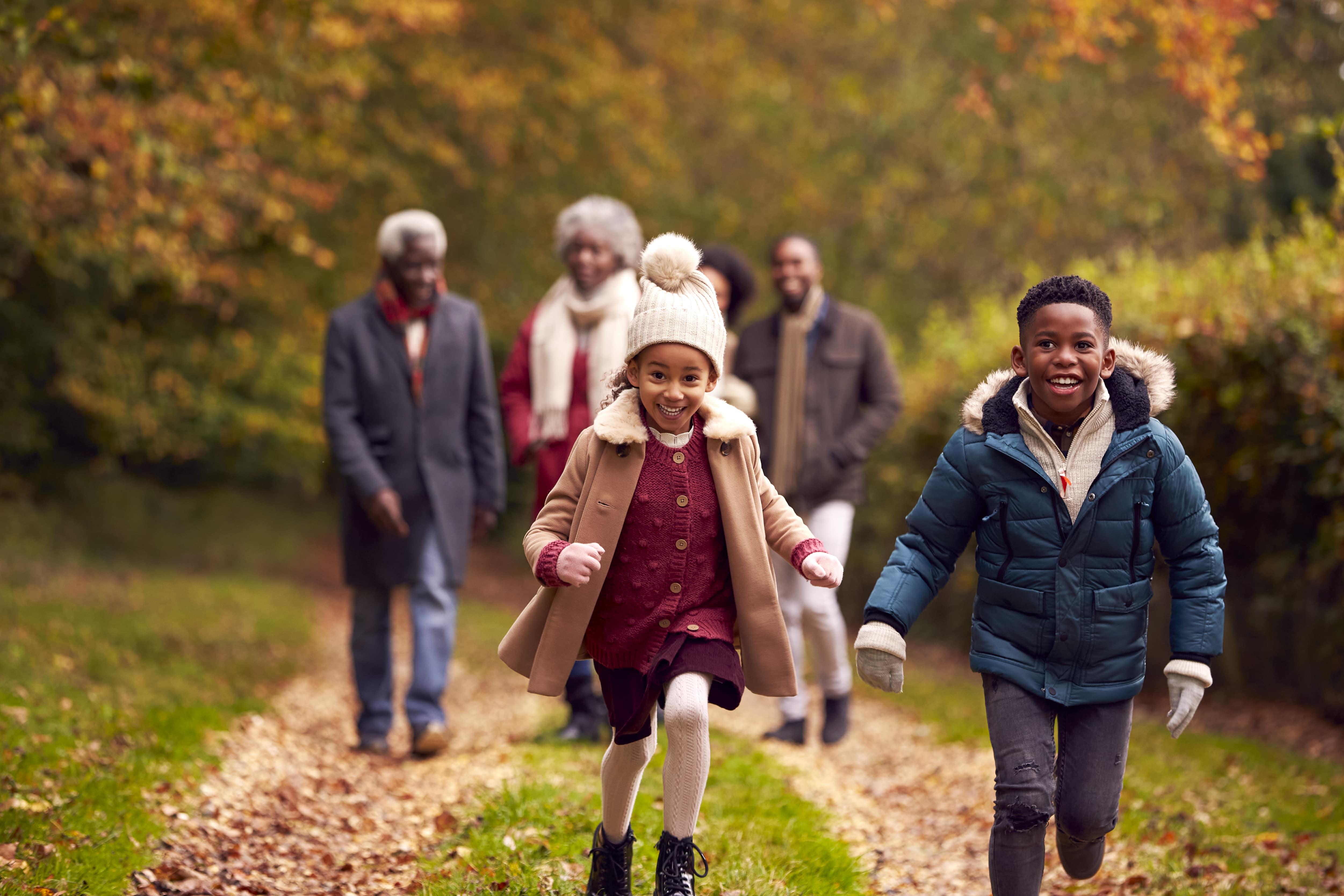 A family enjoying a Fall nature walk. Two young children dressed in Fall jackets run ahead of their parents and grandparents on the hiking trail. 