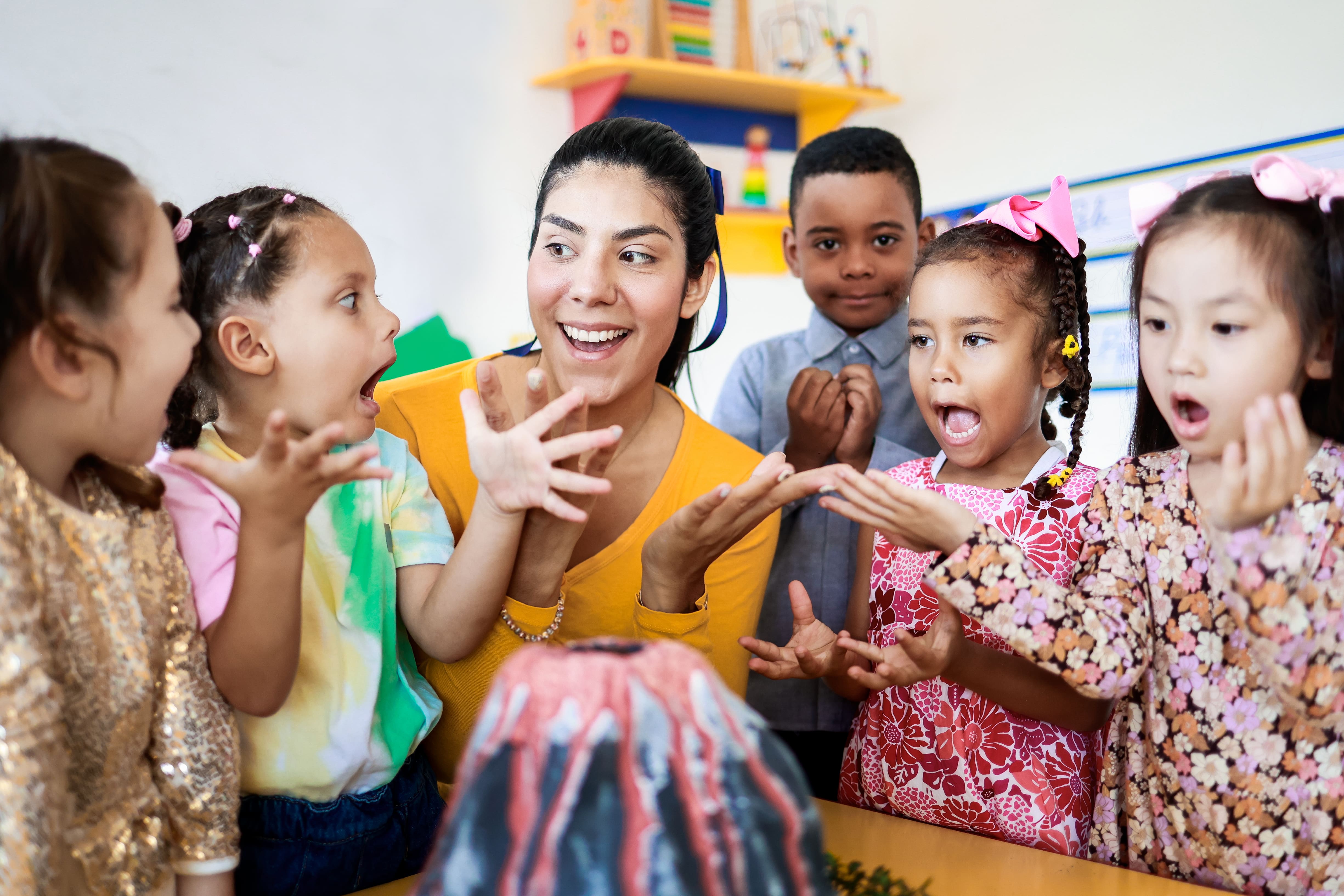 A Preschool educator implementing child-led learning strategies. A young teacher is gathered around a table and a homemade volcano with her many Preschool students. 